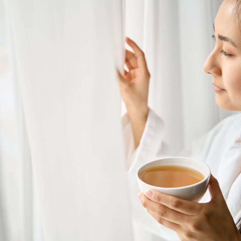 Asian woman moving snow-white tulle on the window to look at the landscape and drinking tea, pleasure morning in luxury hotel room, vacations