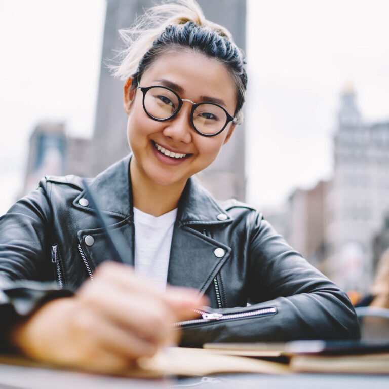 Portrait of happy Chinese tourist sitting in street cafe and smiling at camera enjoying leisure time on vacations, cheerful Japanese hipster girl in trendy spectacles for provide eyes correction