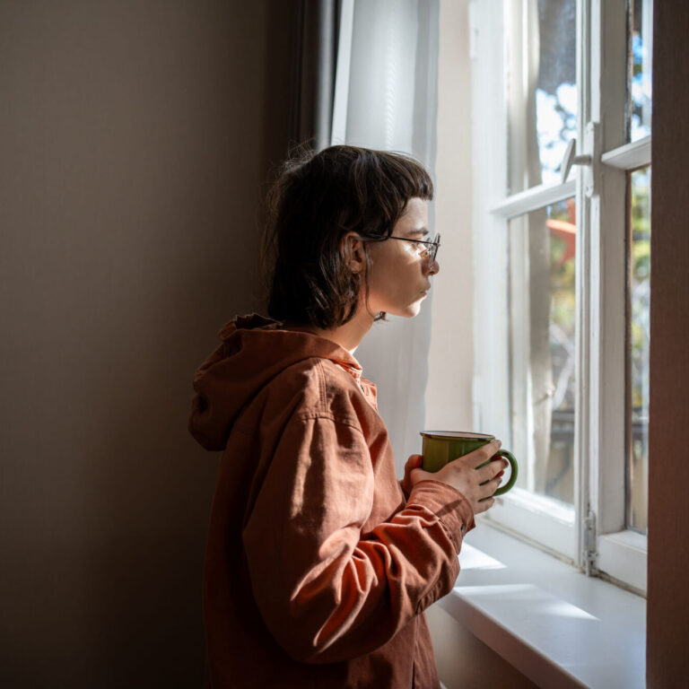 Lonely sad woman with cup of tea looking a window. Unsociable teenage girl spending weekend at home because of friendless, social difficulties, problems in communication, misunderstanding