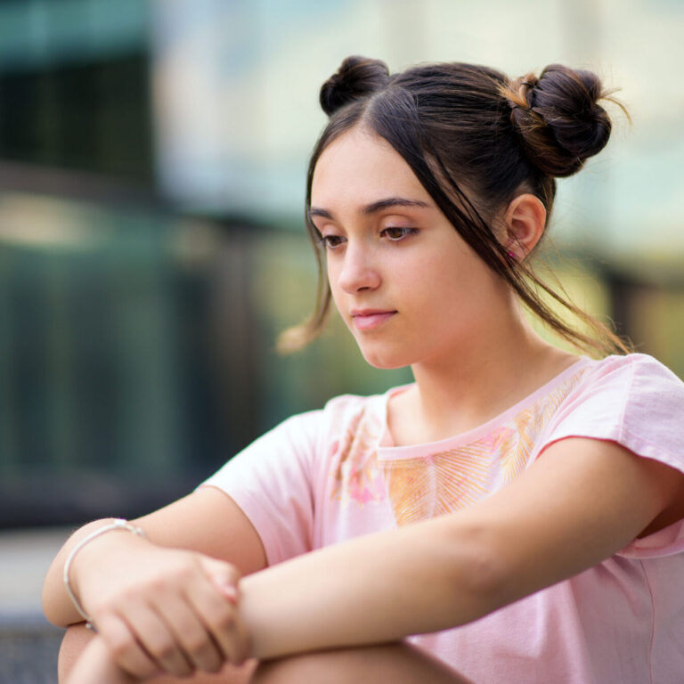 Pensive teenage girl with hair buns sitting alone on street and thinking about life against blurred background
