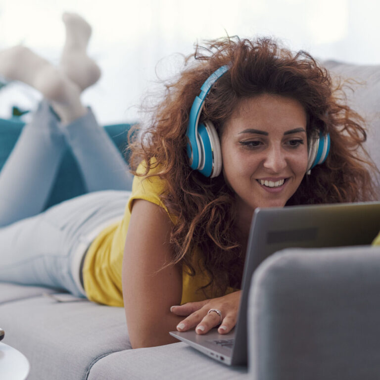 Happy woman relaxing at home, she is lying belly down on the sofa and watching videos on her laptop