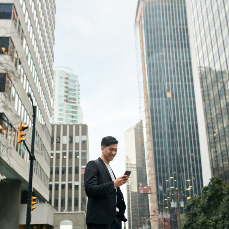 Young happy Asian businessman executive, Japanese business man investor standing in big city street with downtown buildings using cell phone looking at smartphone checking financial apps on mobile.